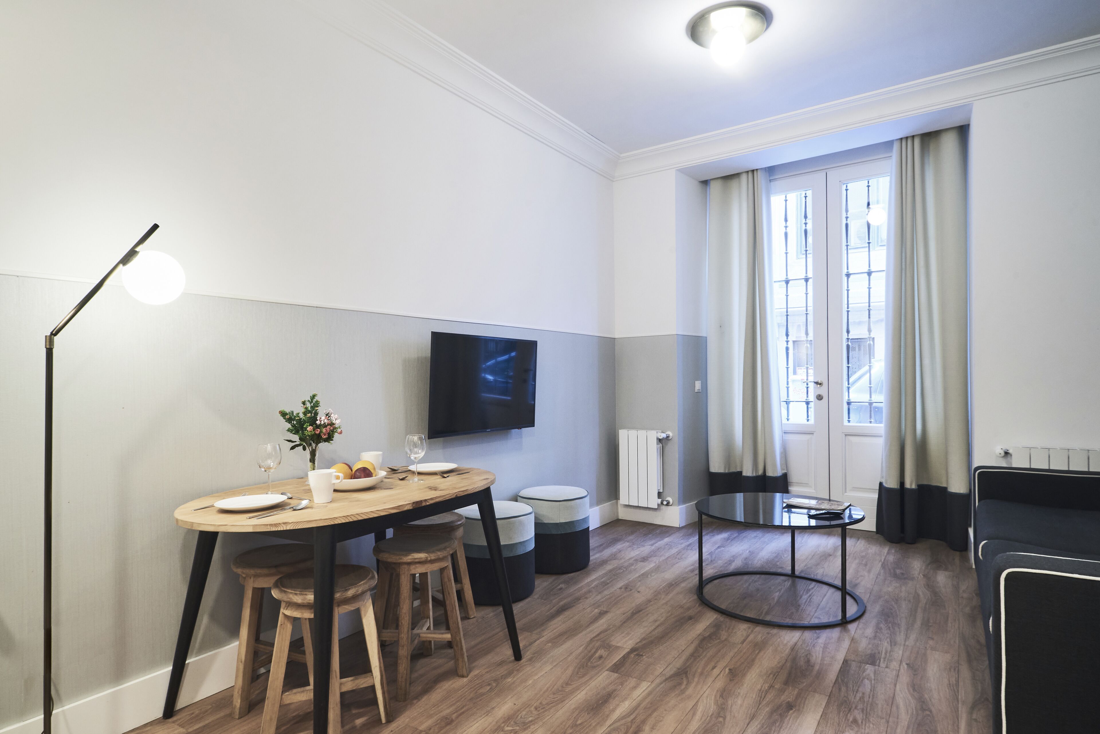 Un salon moderne avec une table à manger en bois, deux tabourets, un téléviseur fixé au mur, une table basse ronde et un canapé noir.