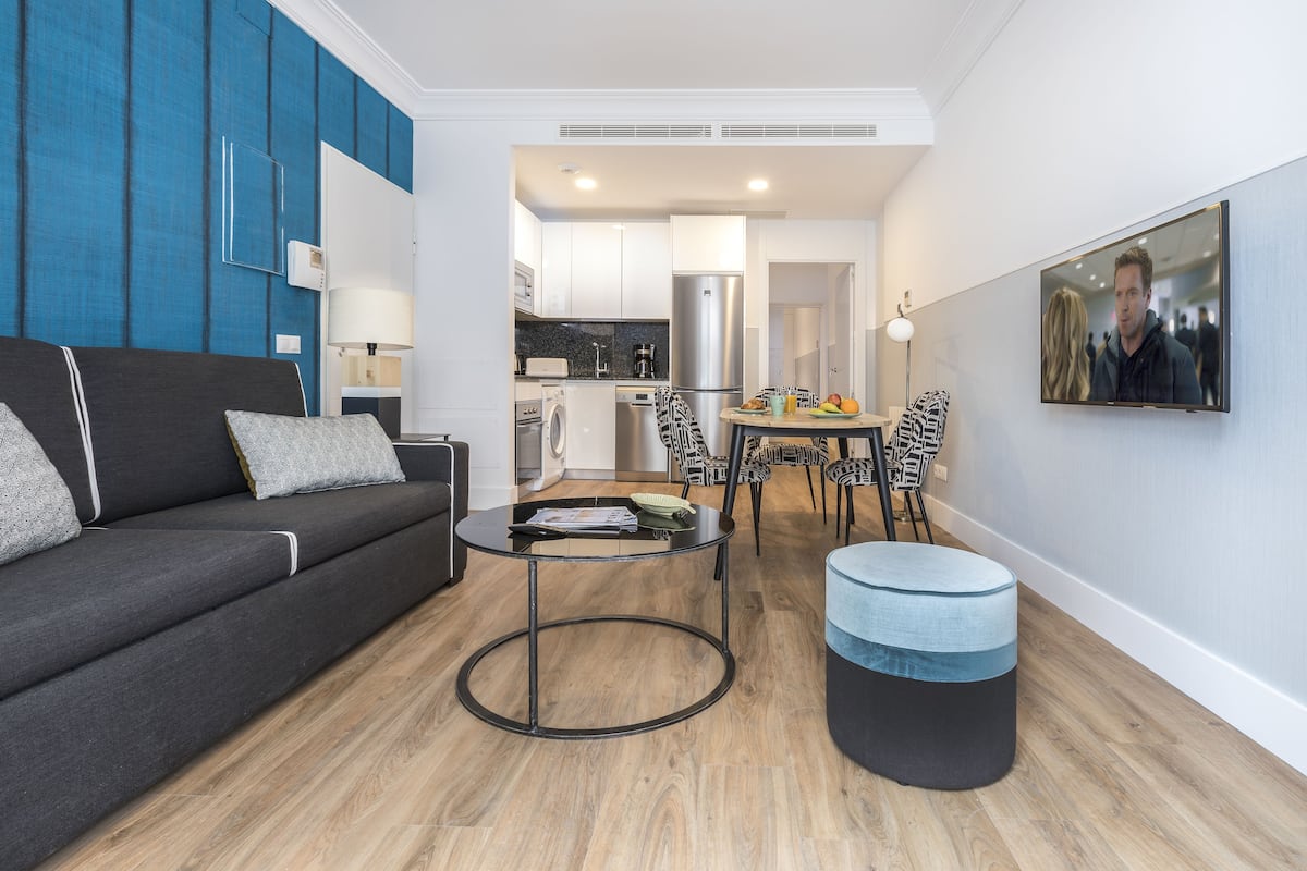 A modern living room with a sofa, a round coffee table, and a round ottoman. There is a kitchen area visible in the background with a dining table and chairs.