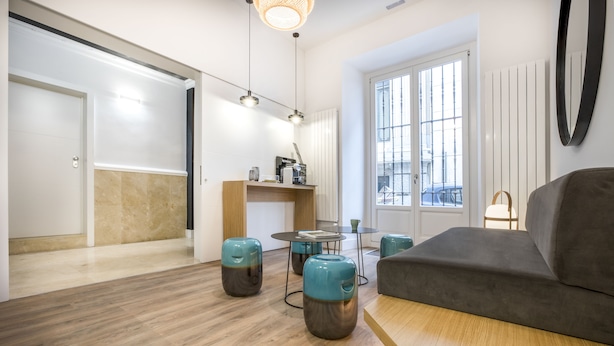 A modern living room with a grey sofa, wooden flooring, and a coffee table. There are two teal stools and a small round table with a coffee machine. - Slow Suites Bellas Artes (Madrid)