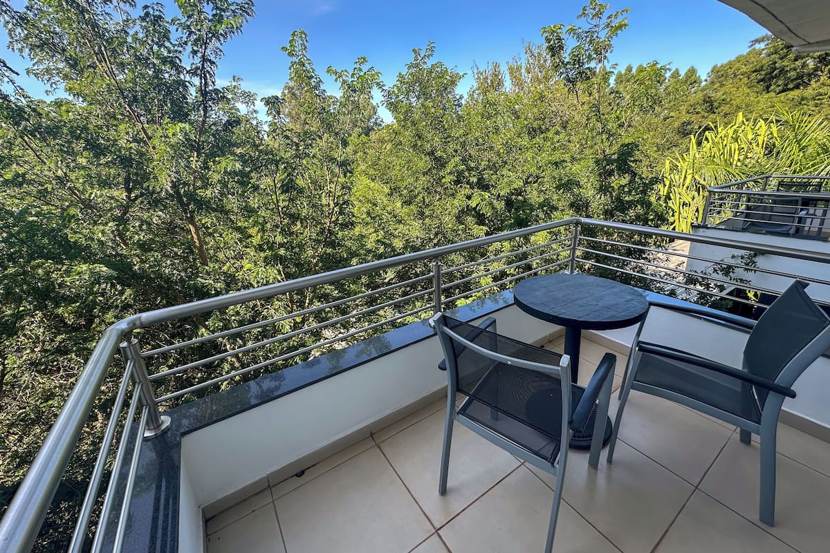 A balcony with a round table and chairs, a metal railing, and a view of dense greenery.