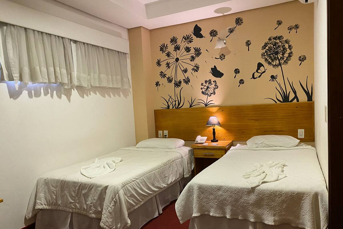 A hotel room with two single beds, a wooden headboard, a nightstand with a lamp, and a wall decoration of flowers and leaves.