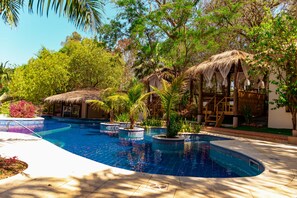 Una piscina en un entorno tropical, rodeada de árboles y una estructura con techo de paja.