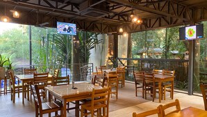 Un restaurante con mesas y sillas de madera, ventanas amplias con follaje en el exterior y dos pantallas mostrando contenido.