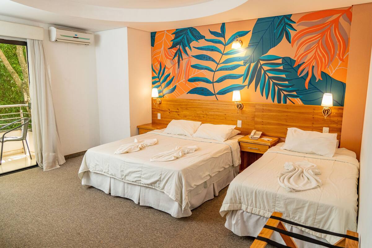 A hotel room with two beds, a wooden headboard, a large tropical leaf mural, and a balcony with a chair.