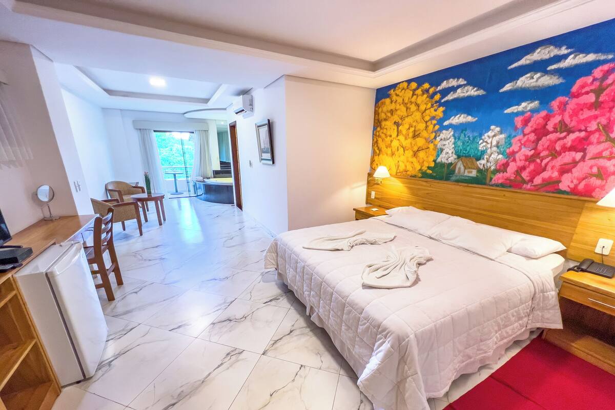 A hotel room with a bed, a desk, a chair, a red rug, and a painting on the wall.