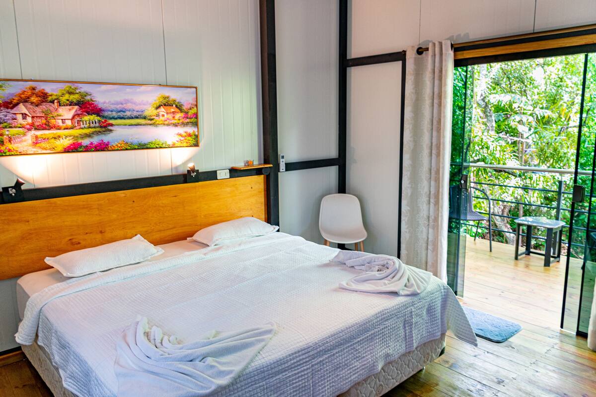 A neatly made bed with white linens and towels, a painting of a landscape on the wall, and a balcony with a view of greenery.
