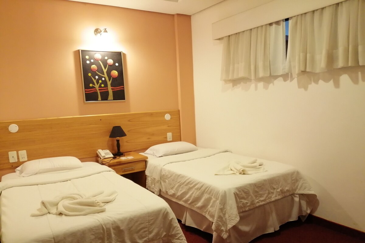 A hotel room with two beds, a nightstand with a lamp, a painting on the wall, and a window with curtains.
