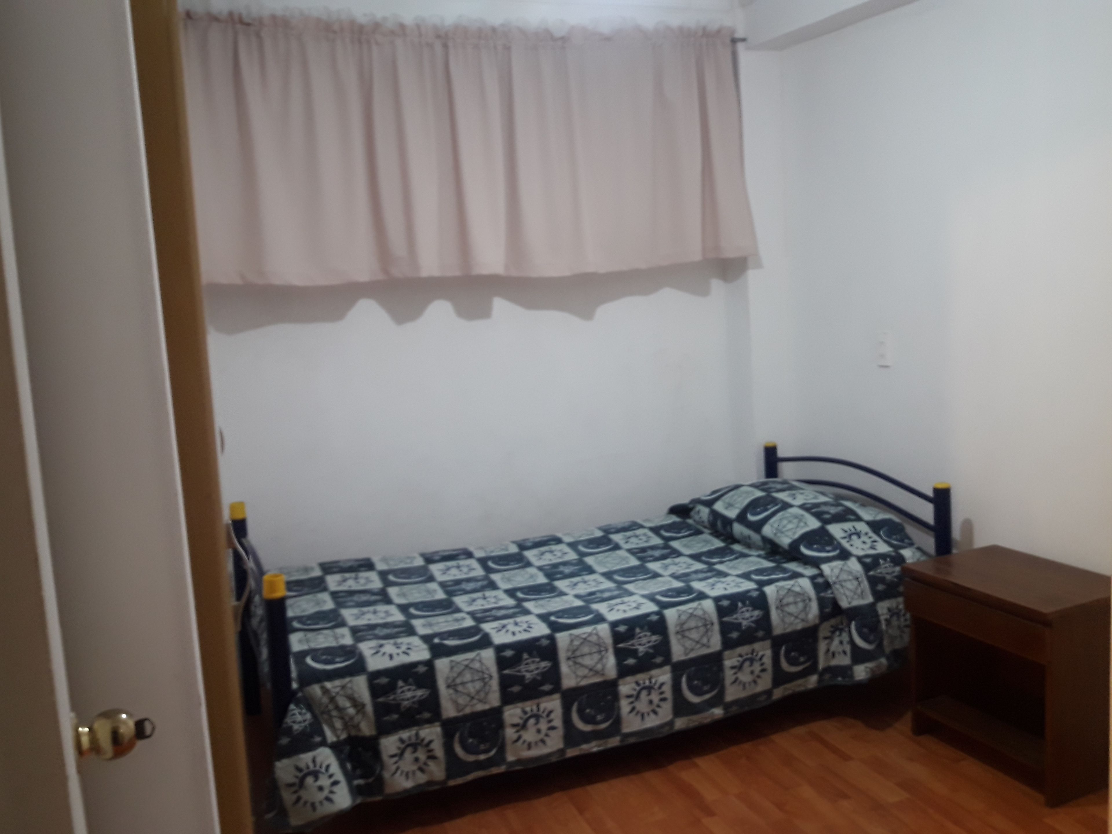 Una habitación pequeña con cama individual, colcha estampada, mesita de noche de madera y ventana con cortina clara.