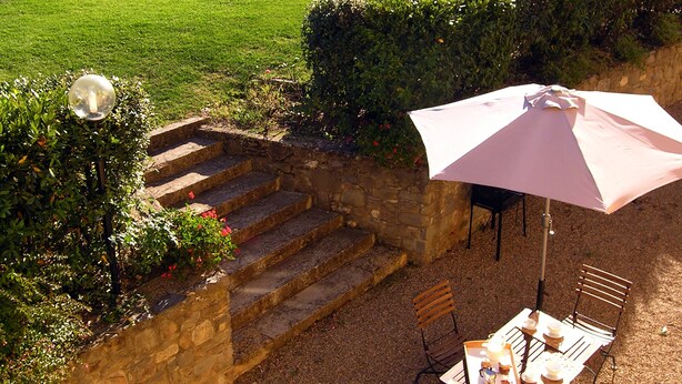 A patio with a table, chairs, and an umbrella. There are steps leading up to the patio, and a stone wall surrounds it. - Agriturismo Il Bellini (Castelfranco Piandisco)