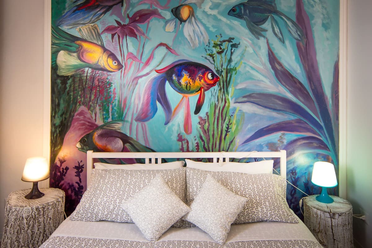 Een slaapkamer met een bed, gestreepte kussens, een nachtkastje met een lamp en een kleurrijke muurschildering.