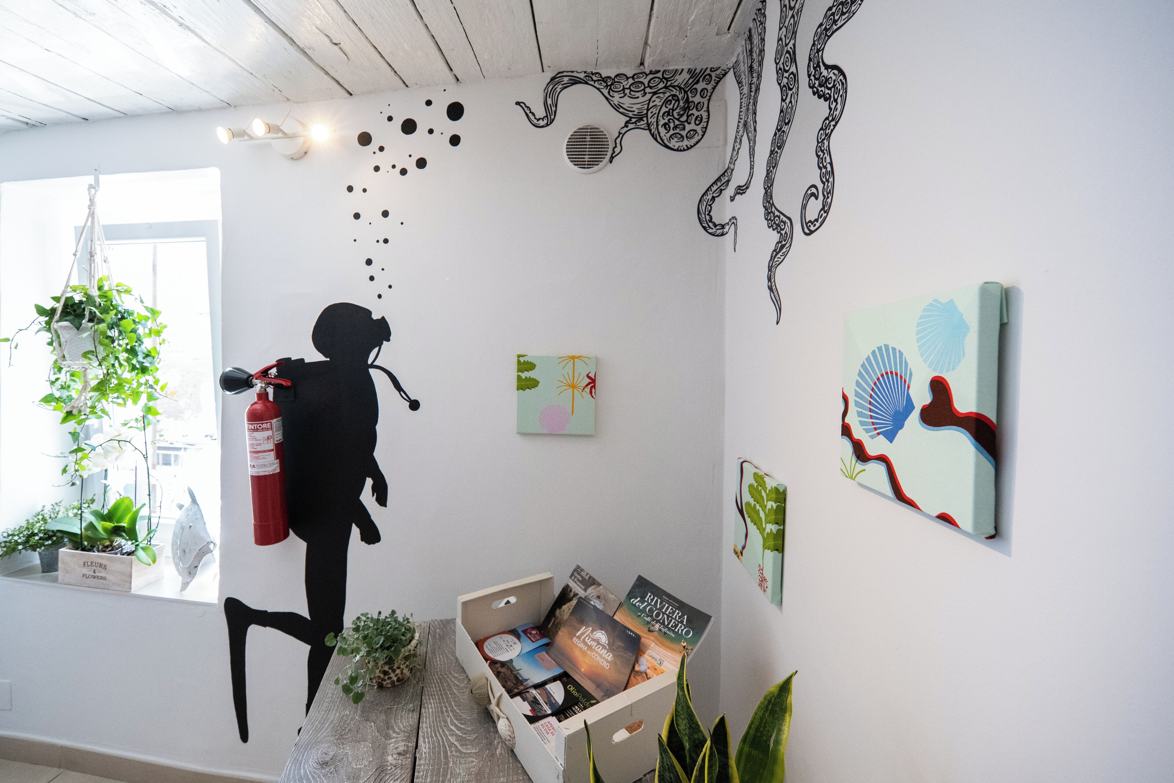 A room with a wooden ceiling, a fire extinguisher, a silhouette decoration, a window with plants, and a box of books.