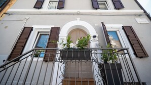 Eine Gebäudefassade mit einem zentralen Bogenportal, braunen Fensterläden und einem Balkon mit Pflanzkübeln.