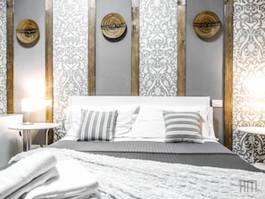 Una cama impecable con almohadas blancas y a rayas, un edredón gris y una pila de toallas blancas. La habitación cuenta con un cabecero de madera, una mesita de noche pequeña con una lámpara y un papel tapiz estampado.