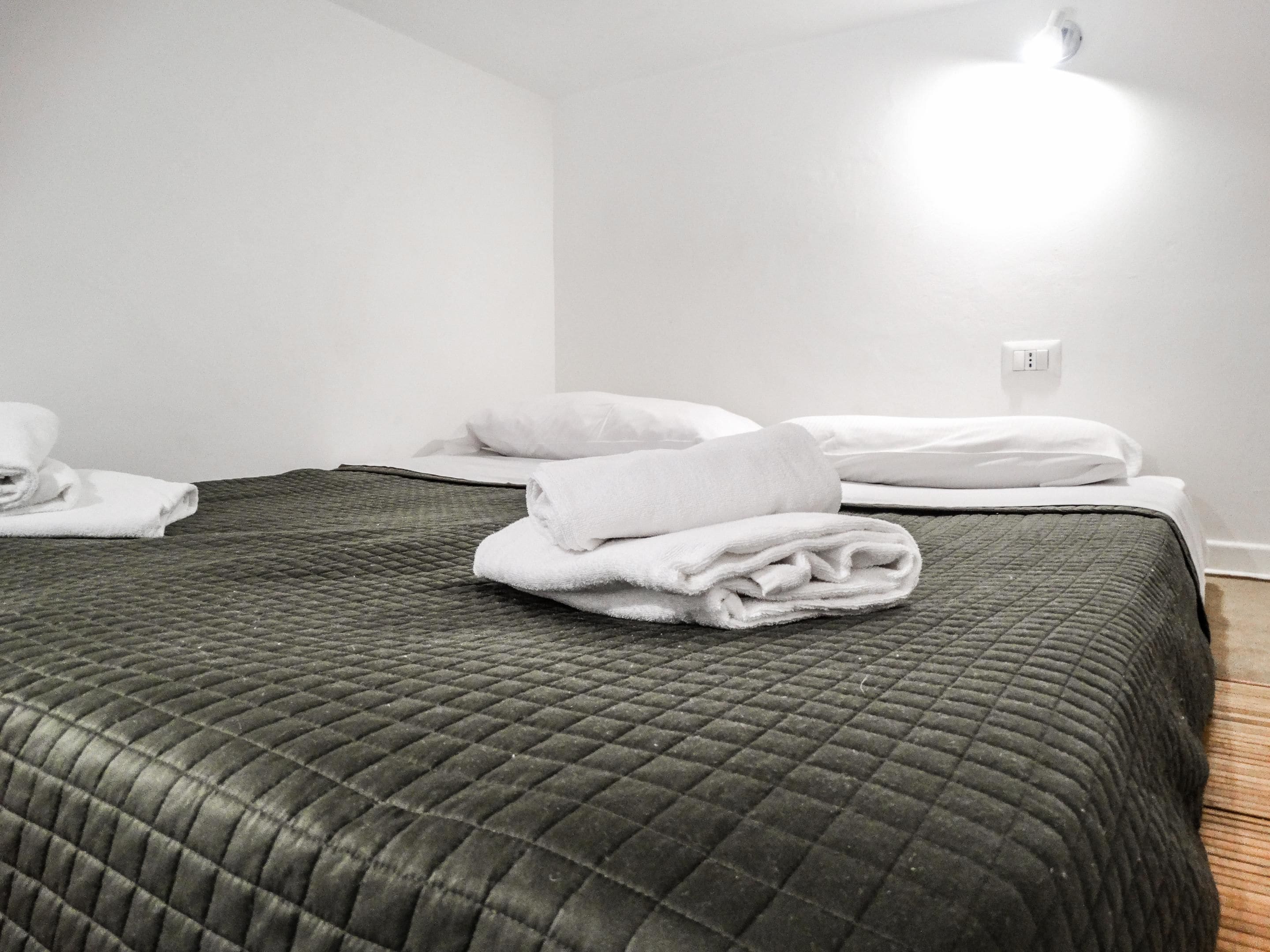 A hotel room with a single bed, a quilted dark green bedspread, and neatly folded white towels on the bed.