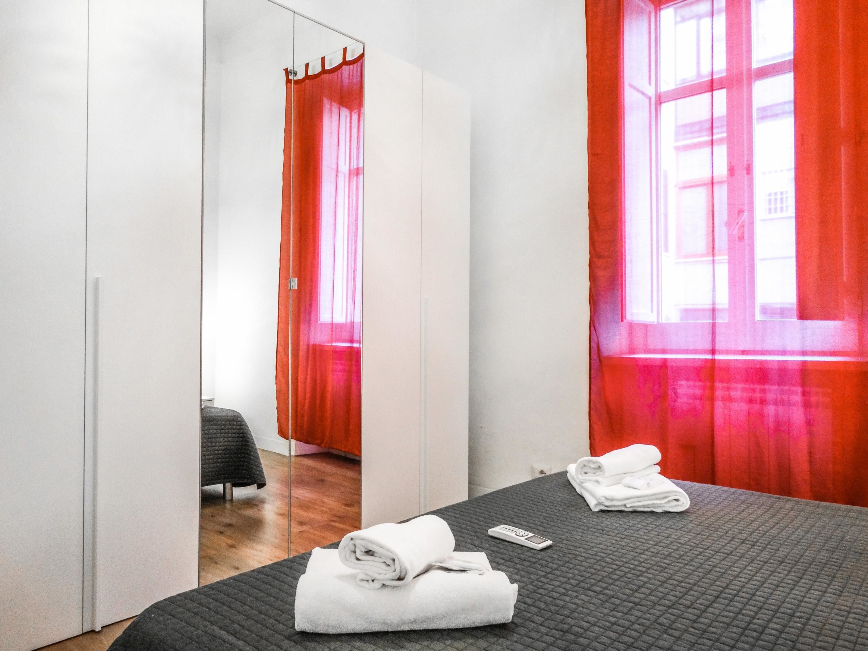 A hotel room with a bed, a mirror, a window with red curtains, and a bedside table with towels and a remote control.