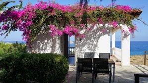 A covered outdoor seating area with a view of the sea, surrounded by lush greenery and vibrant pink flowers. - Bungalow Musuri (Sitia)