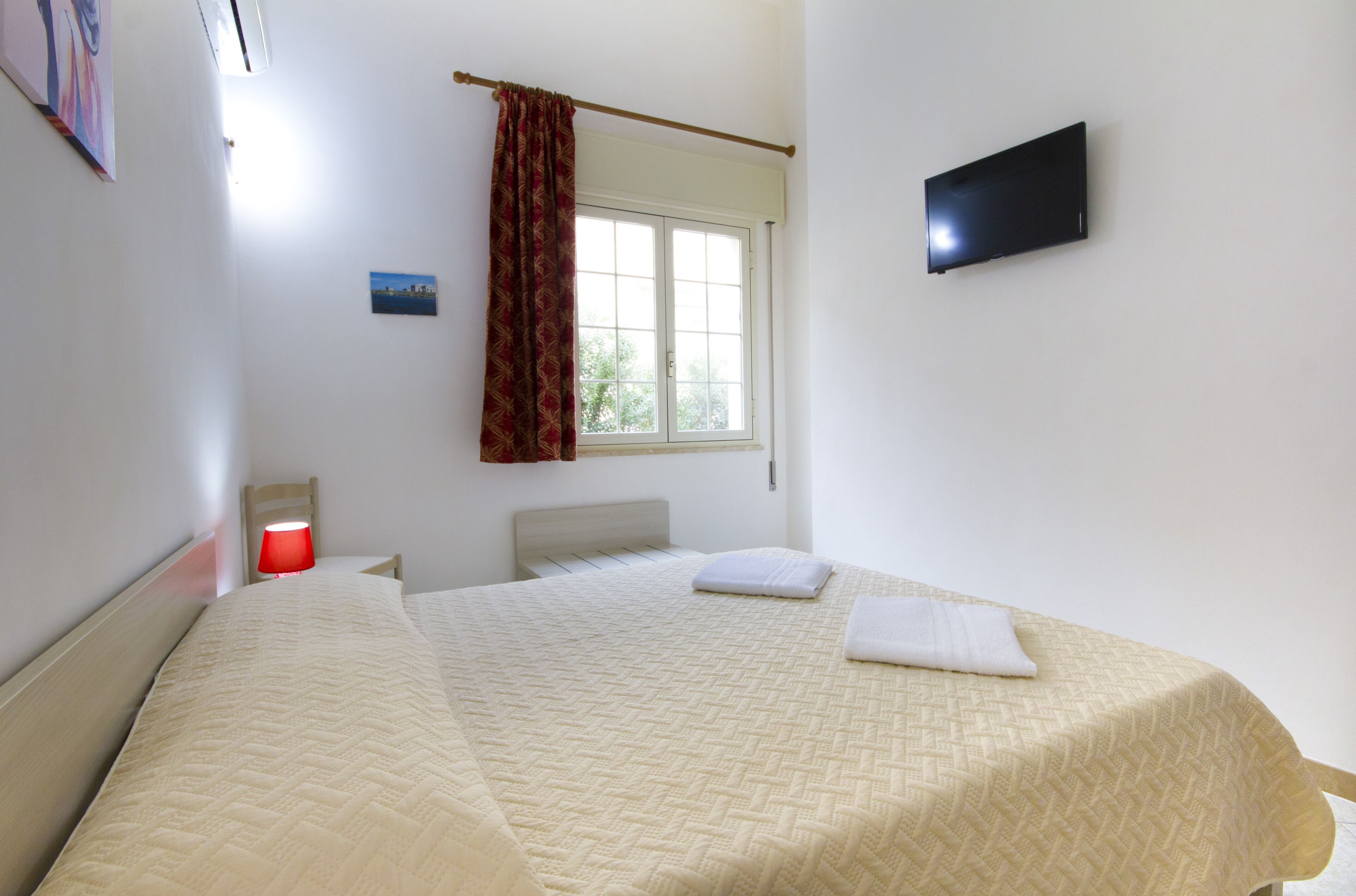 A hotel room with a bed, a window with curtains, a flat-screen TV, and a small table with a lamp.