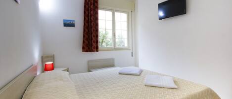 A hotel room with a bed, a window with curtains, a flat-screen TV, and a small table with a lamp.