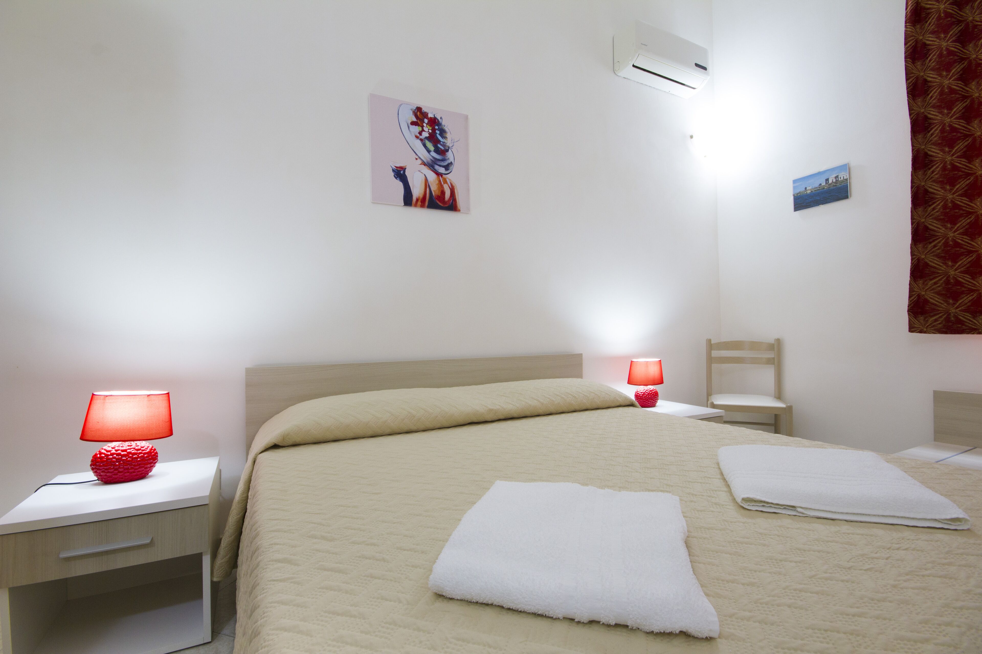 Un letto rifatto con due asciugamani bianchi piegati, una lampada rossa su un comodino, una piccola sedia e un condizionatore d'aria a parete.
