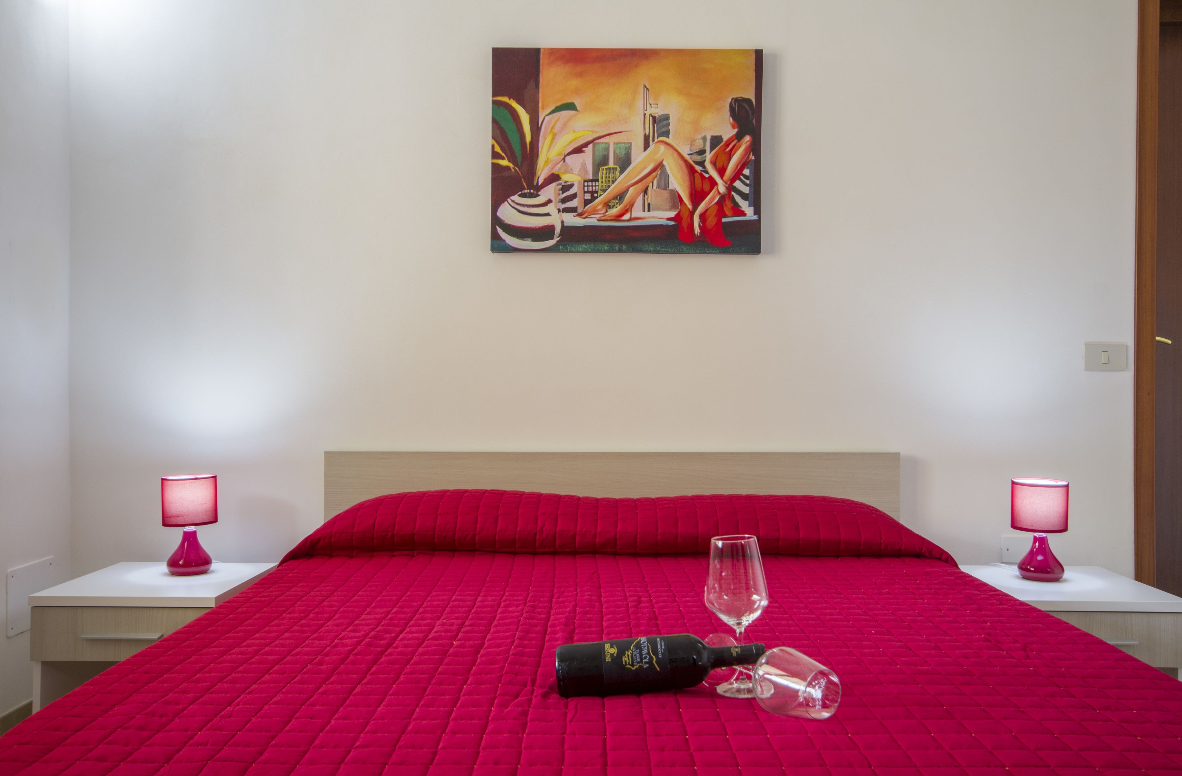 Habitación de hotel con una colcha roja, dos lámparas de noche, una botella y un vaso sobre la cama, y un cuadro en la pared.