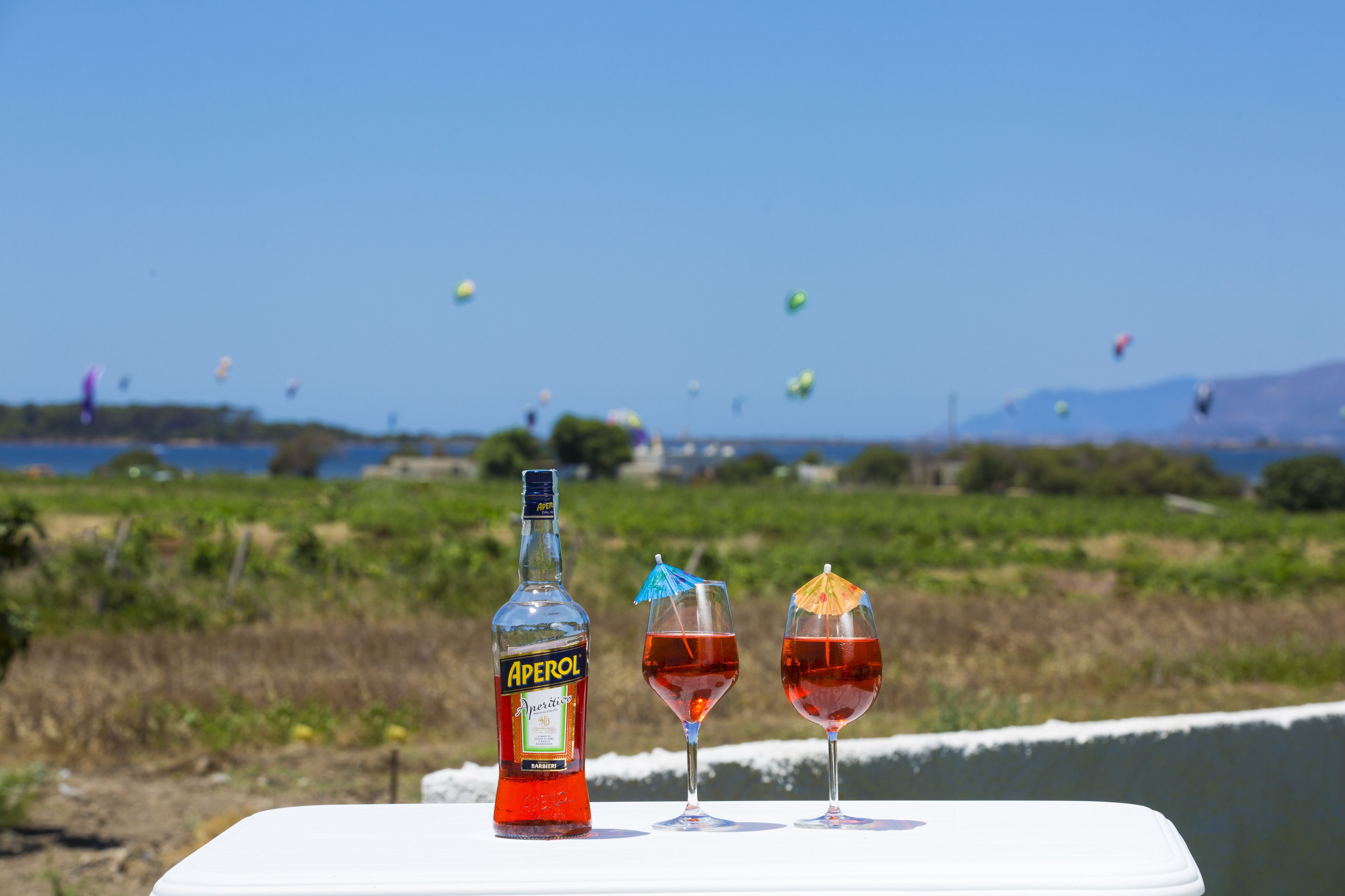 A bottle of Aperol and two glasses of red drink on a white table with a scenic view of kites in the background.