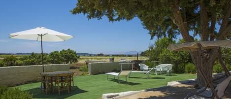 Um pátio externo com mesa e cadeiras, um guarda-sol branco e vista para o campo.