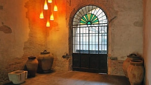 Um quarto rústico com parede de tijolos, uma ampla janela em arco com desenho em vitral verde e amarelo, e vários vasos de barro grandes.