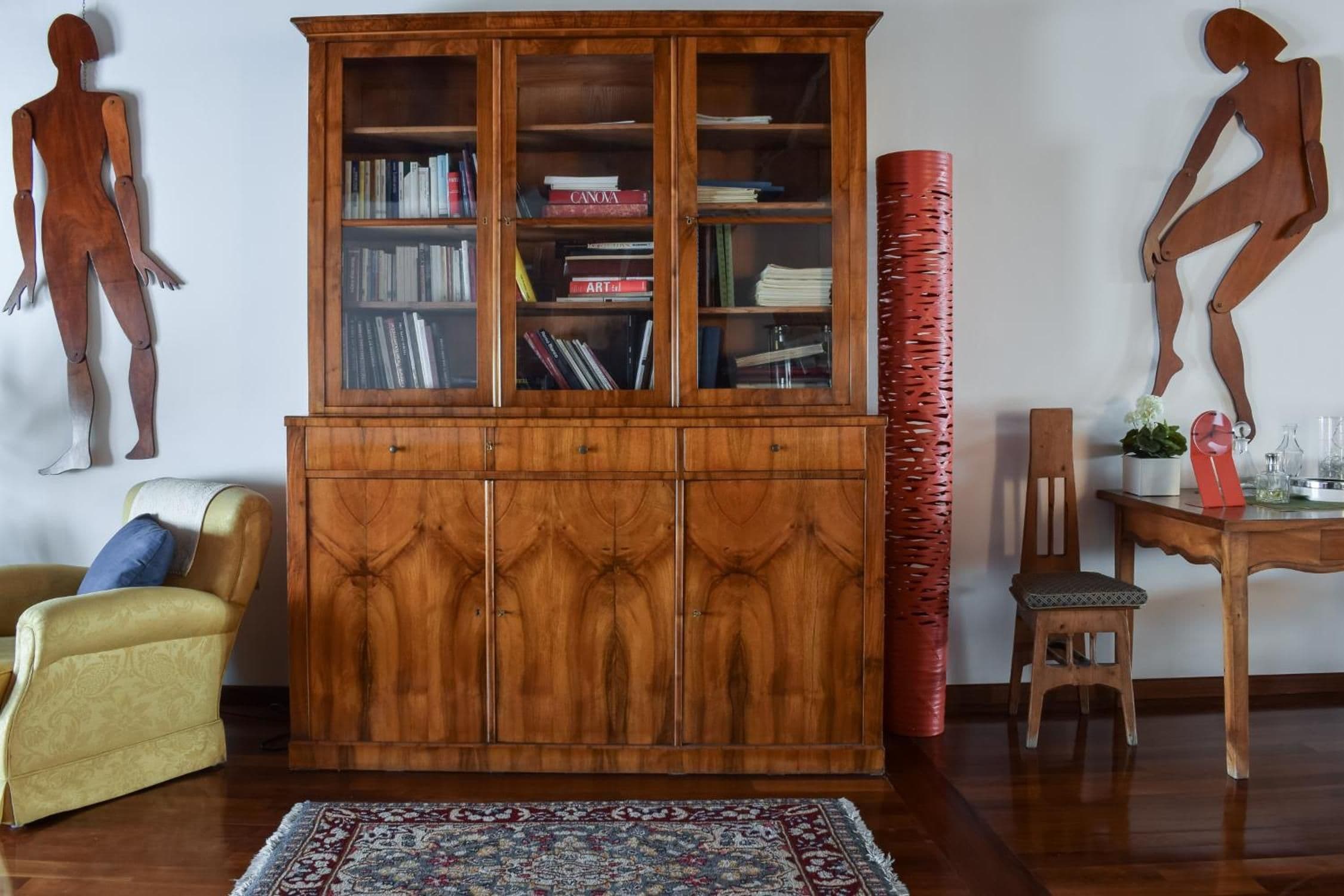 A room with a wooden cabinet, a patterned rug, a yellow armchair, a dining table with a red chair, and two wall-mounted sculptures.