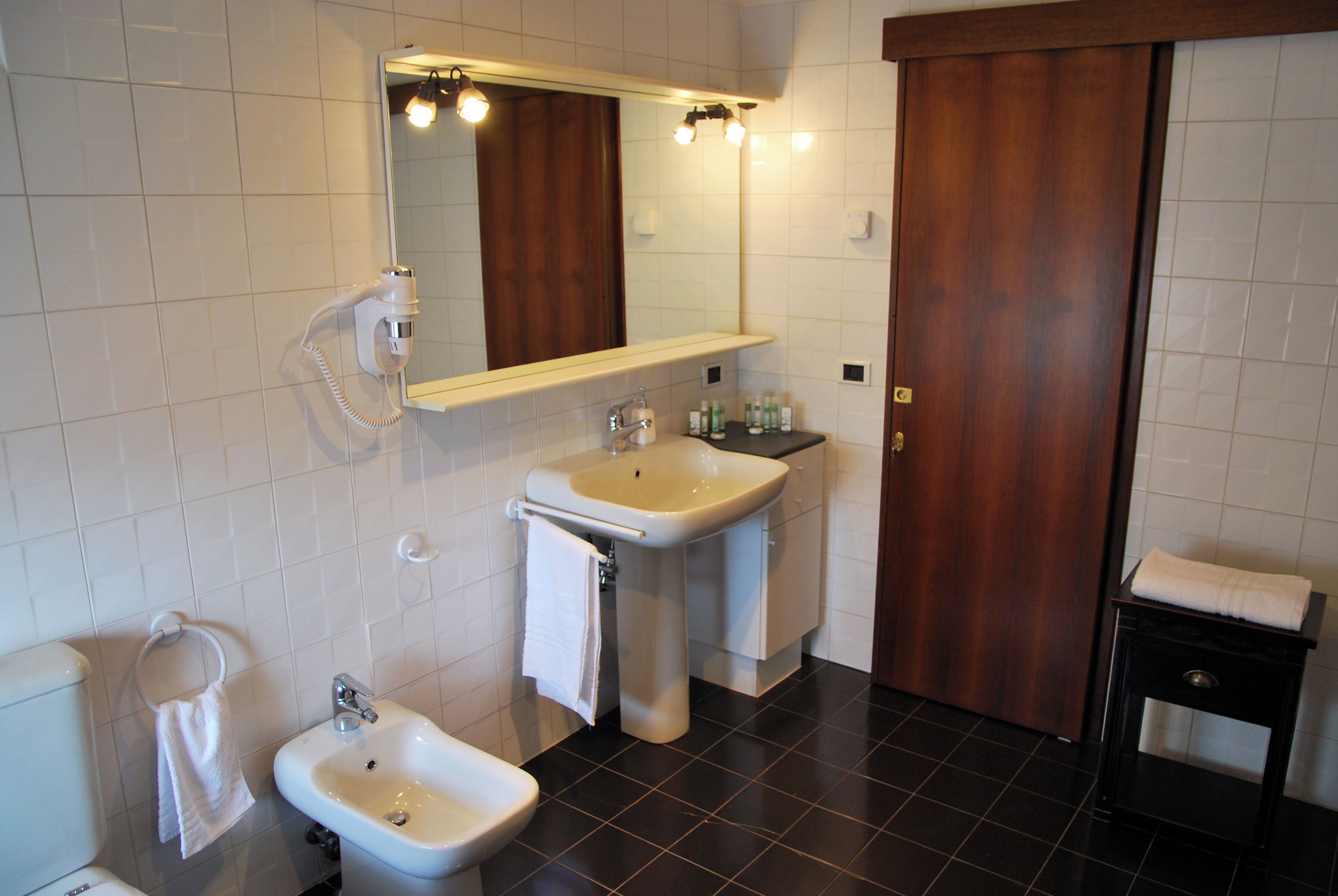 A bathroom with a sink, bidet, and mirror. There is a towel rack and a wooden door.