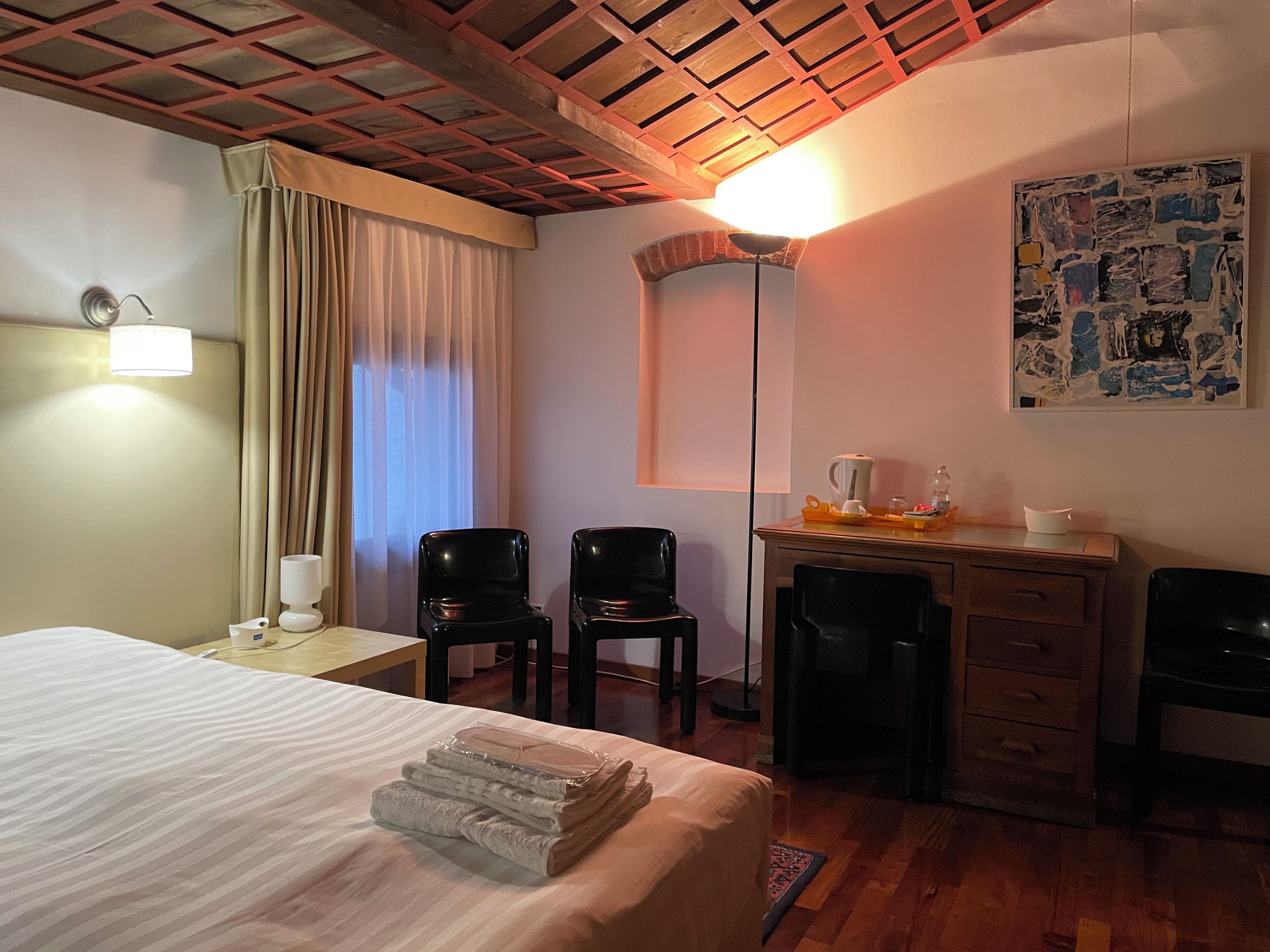 A hotel room with a bed, two chairs, a desk with a lamp, a painting on the wall, and a window with curtains.