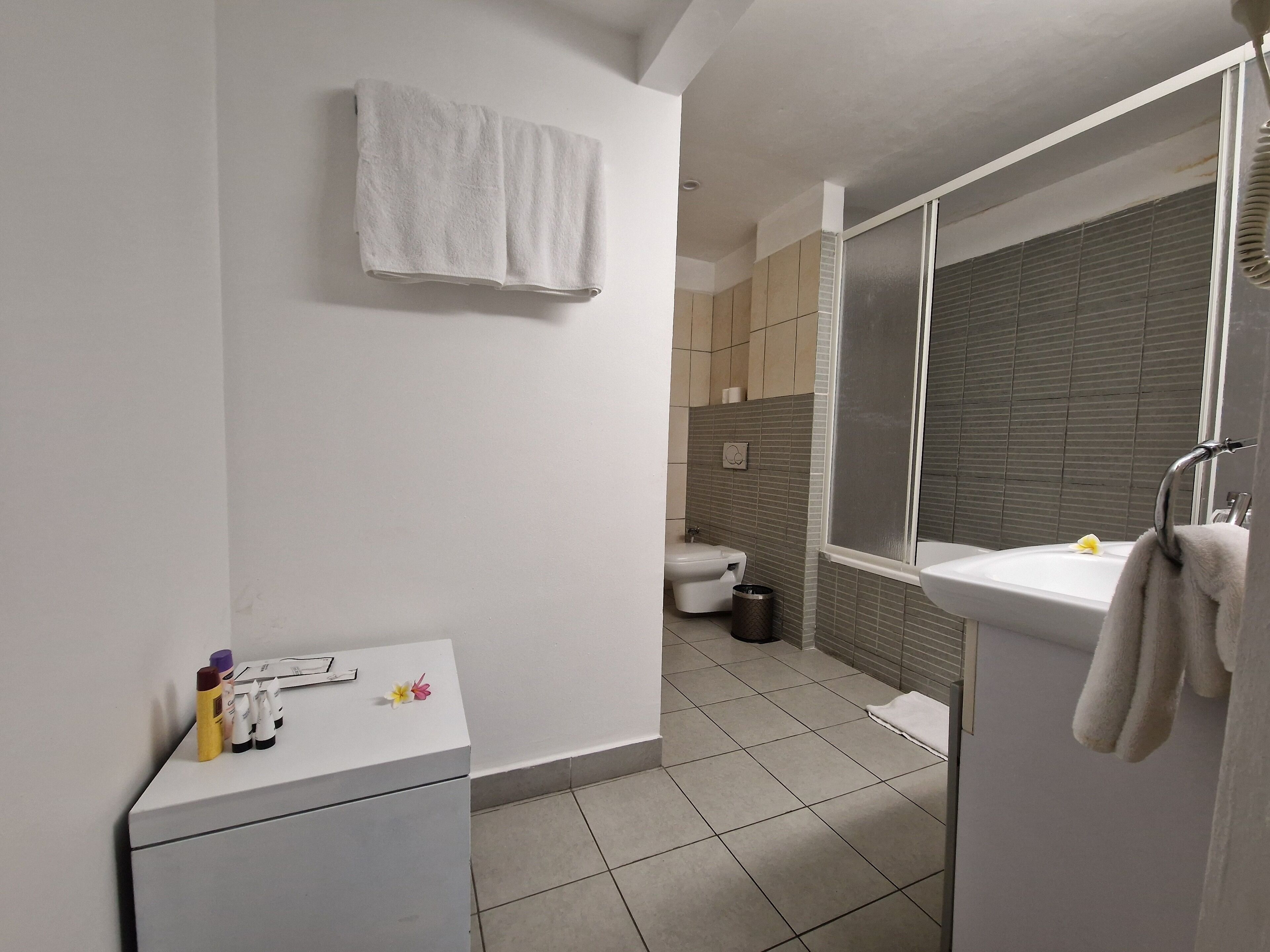 A modern bathroom with a white sink, a mirror, and a towel hanging on the wall. There is a toilet and a shower area visible in the background.