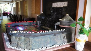 A stone fountain with water cascading into a pond, surrounded by white pebbles and red flowers. -  Itsandra Beach Hotel & Resort (Batsa Itsandra)