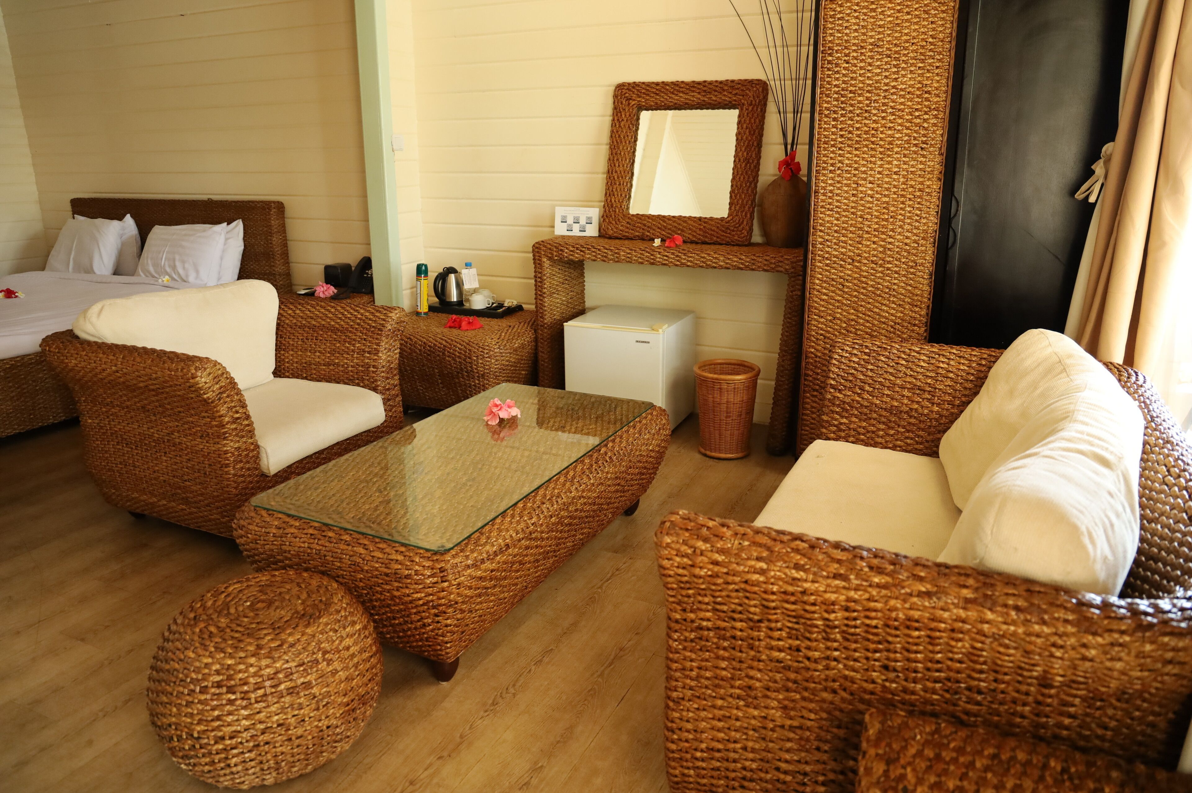 A room with wicker furniture, a glass-top coffee table, a bed, a small refrigerator, and a mirror.