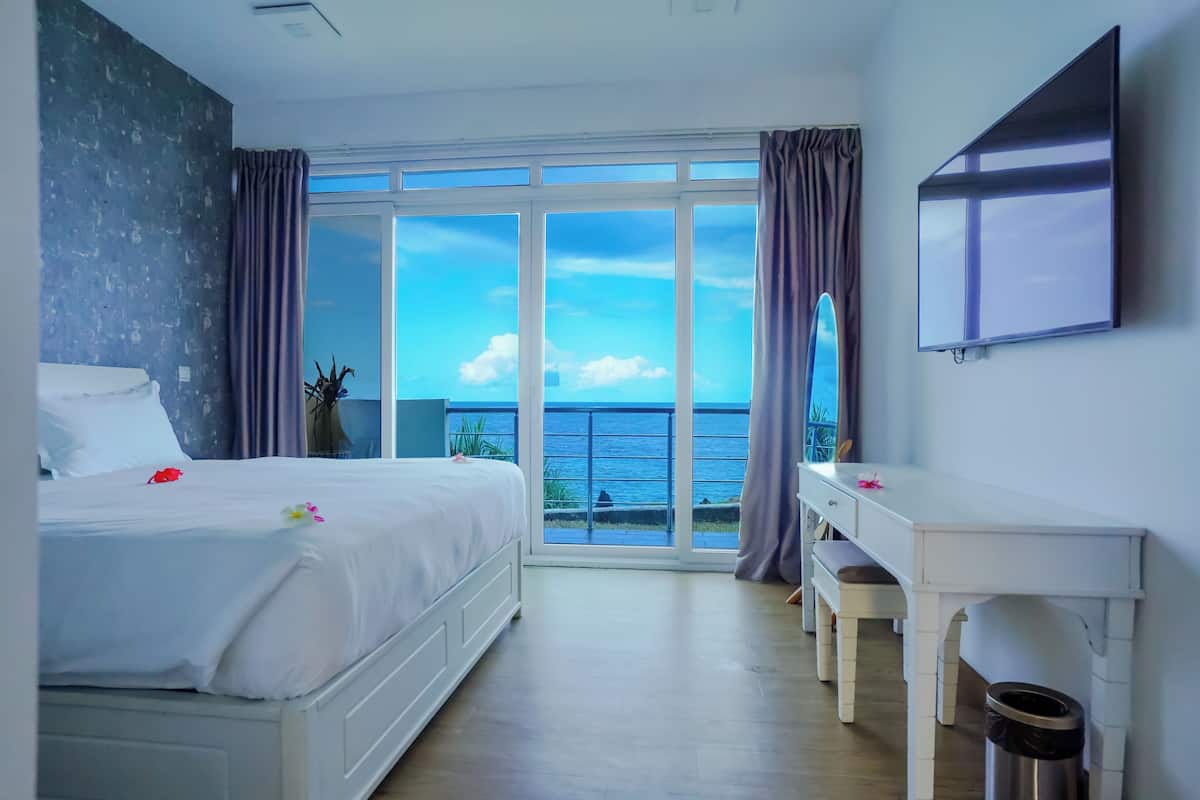 Quarto de hotel com cama, escrivaninha, televisão e uma grande janela com vista para o mar.