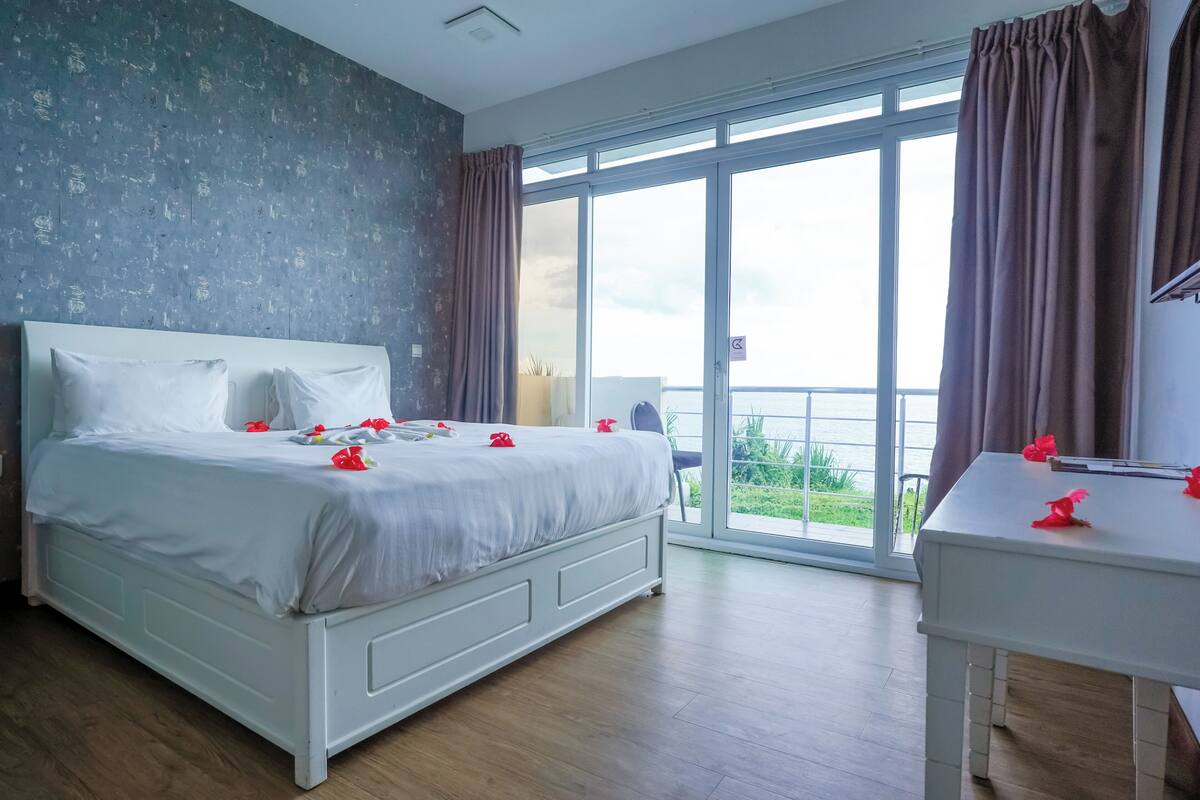 Quarto com uma cama grande, uma escrivaninha branca e vista para o exterior através de uma janela ampla.