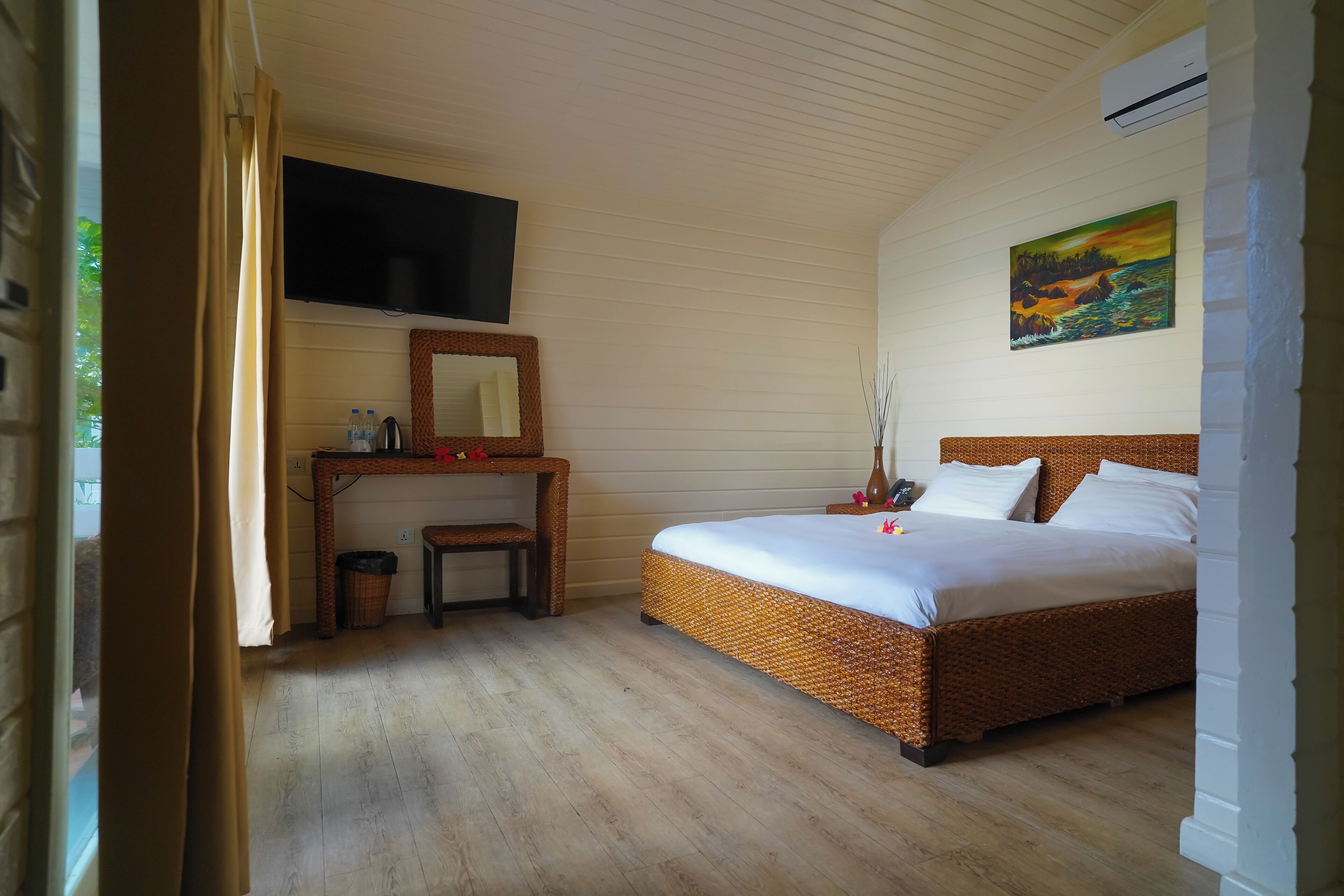 Un dormitorio con suelo de madera, una cama, un televisor montado en la pared, un cuadro y una pequeña mesa con un espejo.