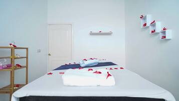 Un lit bien fait, recouvert d’une courtepointe blanche et bleue, avec une serviette enroulée et des pétales de rose rouge éparpillés dessus. Une étagère en bois, sur laquelle sont posés divers objets, se trouve à gauche, et une porte blanche est visible au fond.