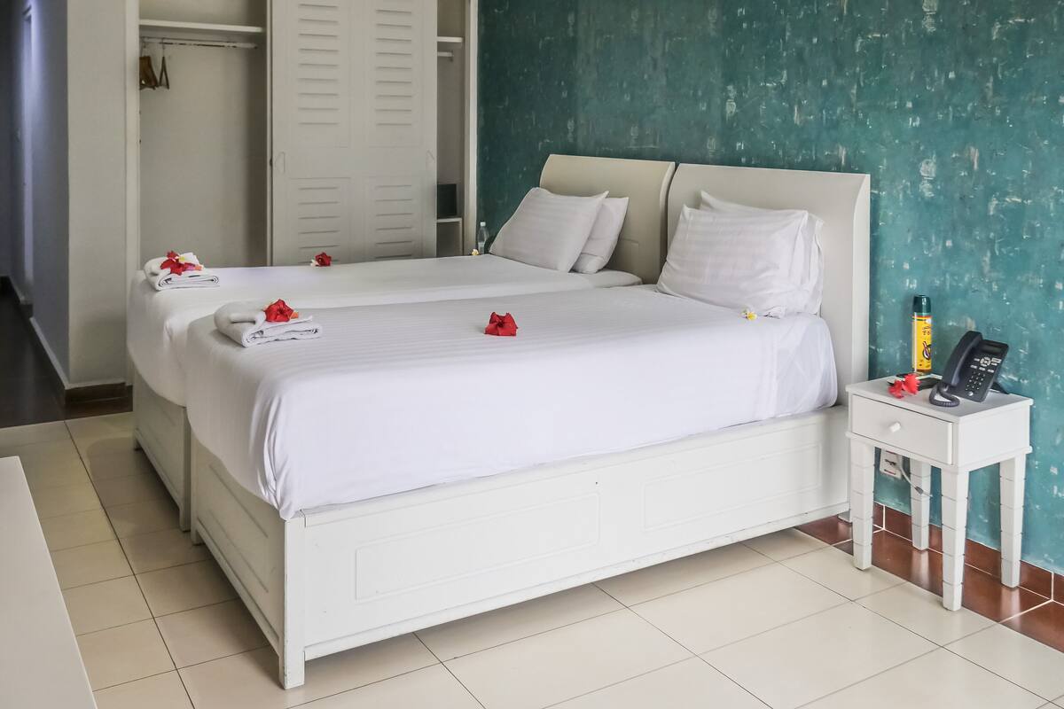 Quarto de hotel com duas camas, roupa de cama branca, mesa de cabeceira com telefone e garrafa, e parede cor de verde-água.