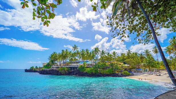 A beach with clear turquoise water, palm trees, and a resort with buildings and a beach area. -  Itsandra Beach Hotel & Resort (Batsa Itsandra)
