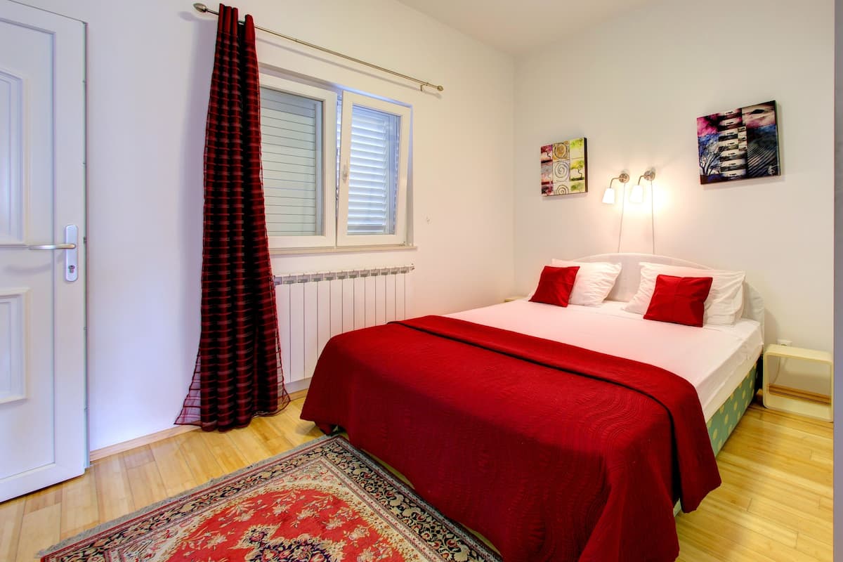 A bedroom with a bed, a red and white color scheme, a radiator, a window with blinds, a door, and a wall-mounted artwork.