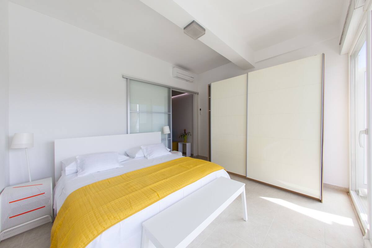 A modern bedroom with a large bed, a bedside table, a bench, a wardrobe, and a sliding door.