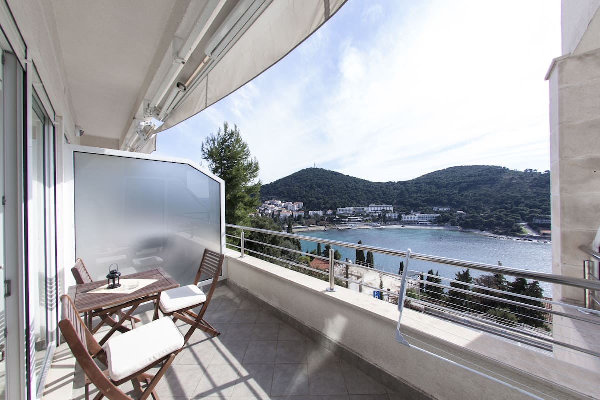 A balcony with a table and chairs, overlooking a body of water and a hillside.