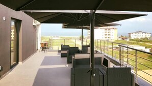 A balcony with outdoor furniture, a large umbrella, and a view of the sea and buildings.