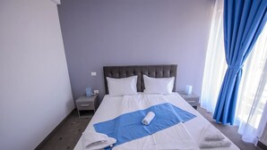 A neatly made bed with white linens, a blue blanket, and rolled towels. There is a nightstand with a lamp and a power outlet. The room has a grey headboard and a window with blue curtains.