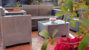 An outdoor patio area with wicker furniture, including sofas and a table, surrounded by greenery and a tiled floor. - B&B Villa Hirschen (Zafferana Etnea)