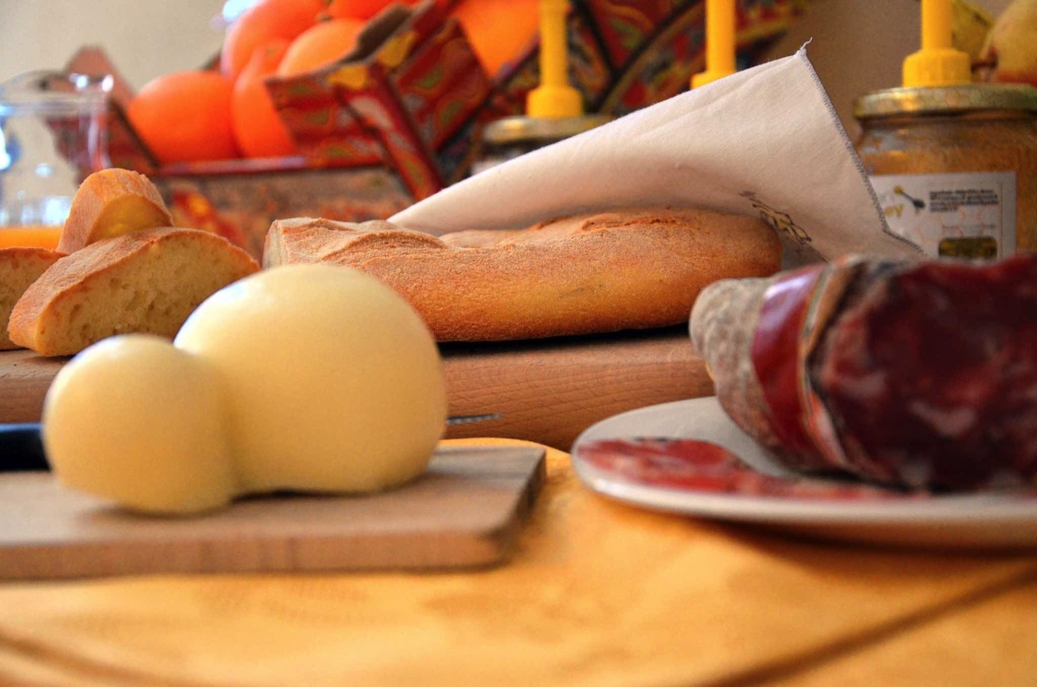 A wooden cutting board with a loaf of bread, a whole cheese, and a sliced ham on a table.
