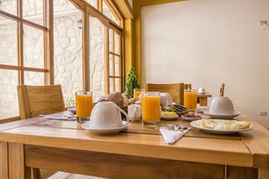Una mesa de desayuno con diversos alimentos y bebidas, en una habitación con ventanas amplias y pared de piedra.