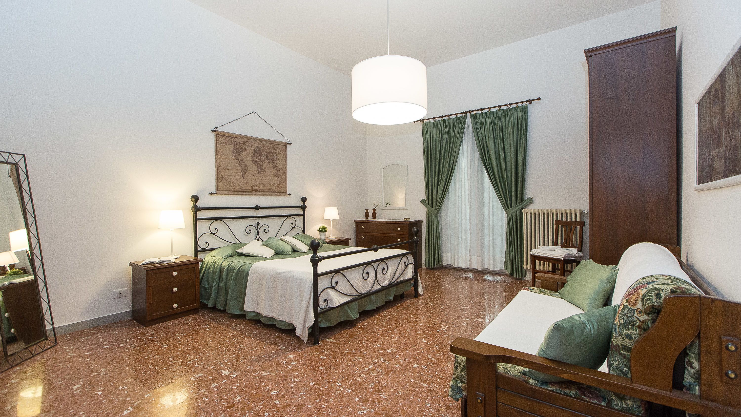 A bedroom with a bed, bedside tables, a dresser, a chair, a hanging light fixture, a map on the wall, and a radiator.