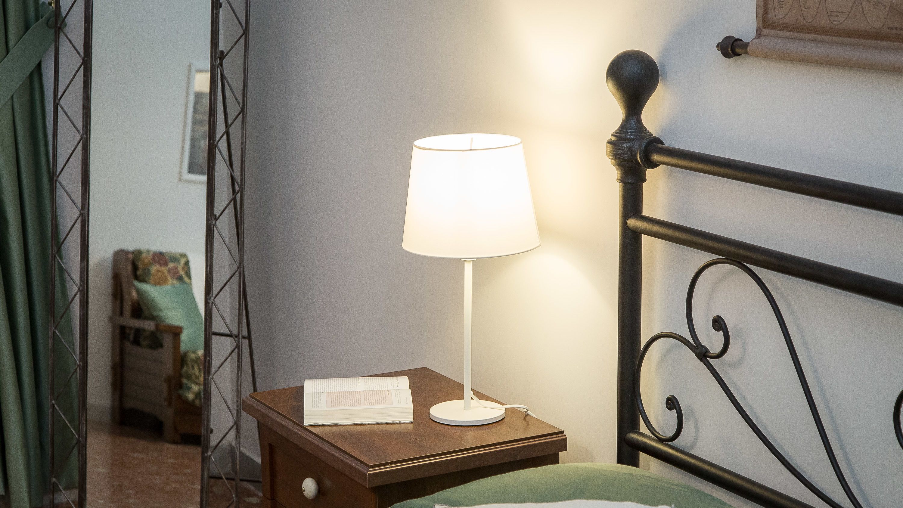 A neatly made bed with a green blanket, a bedside table with a lamp, and a metal headboard.