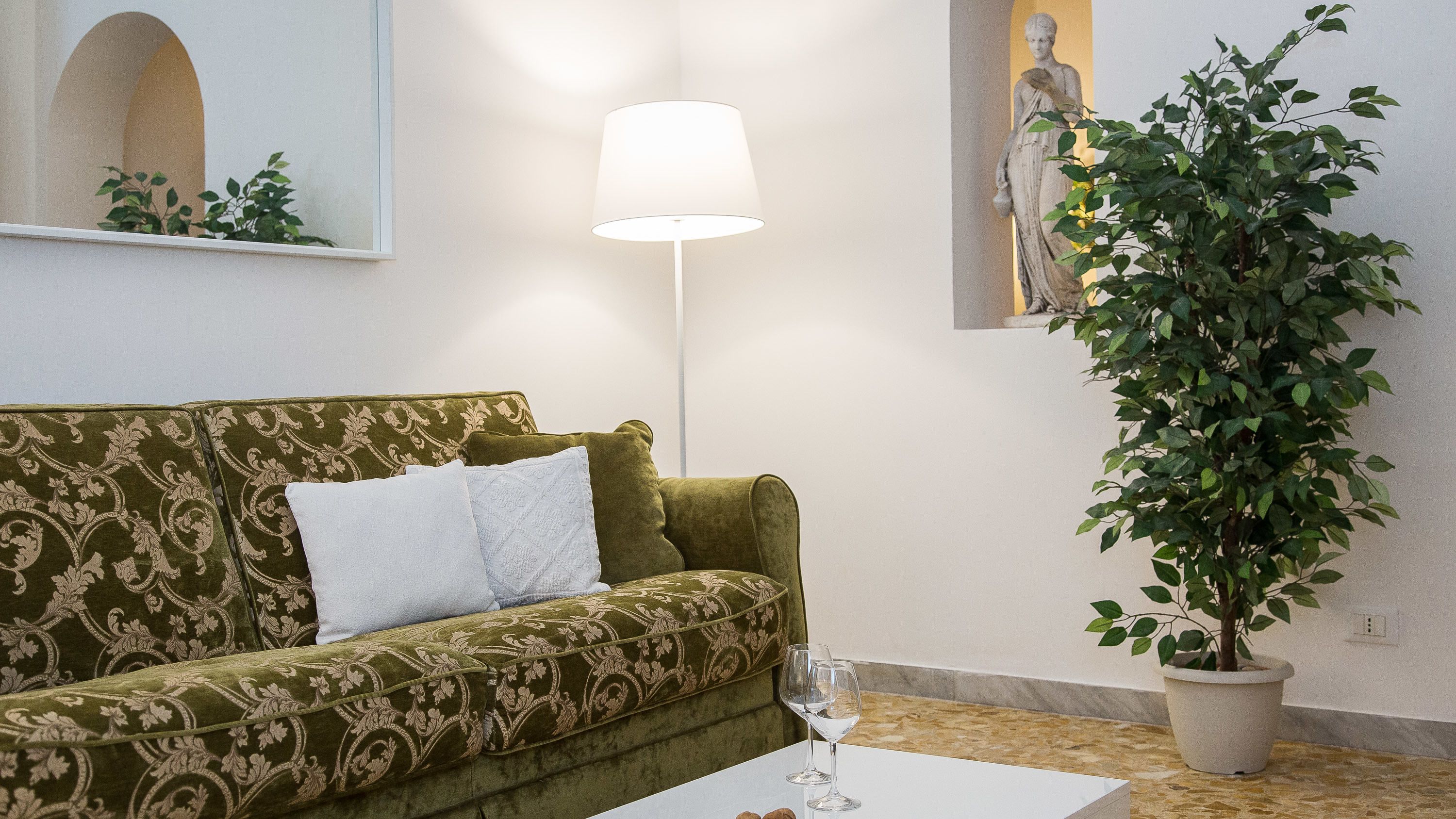 Ein Wohnzimmer mit einem grün gemusterten Sofa, einem weißen Tisch mit einem Weinglas, einer Stehlampe und einer Topfpflanze.